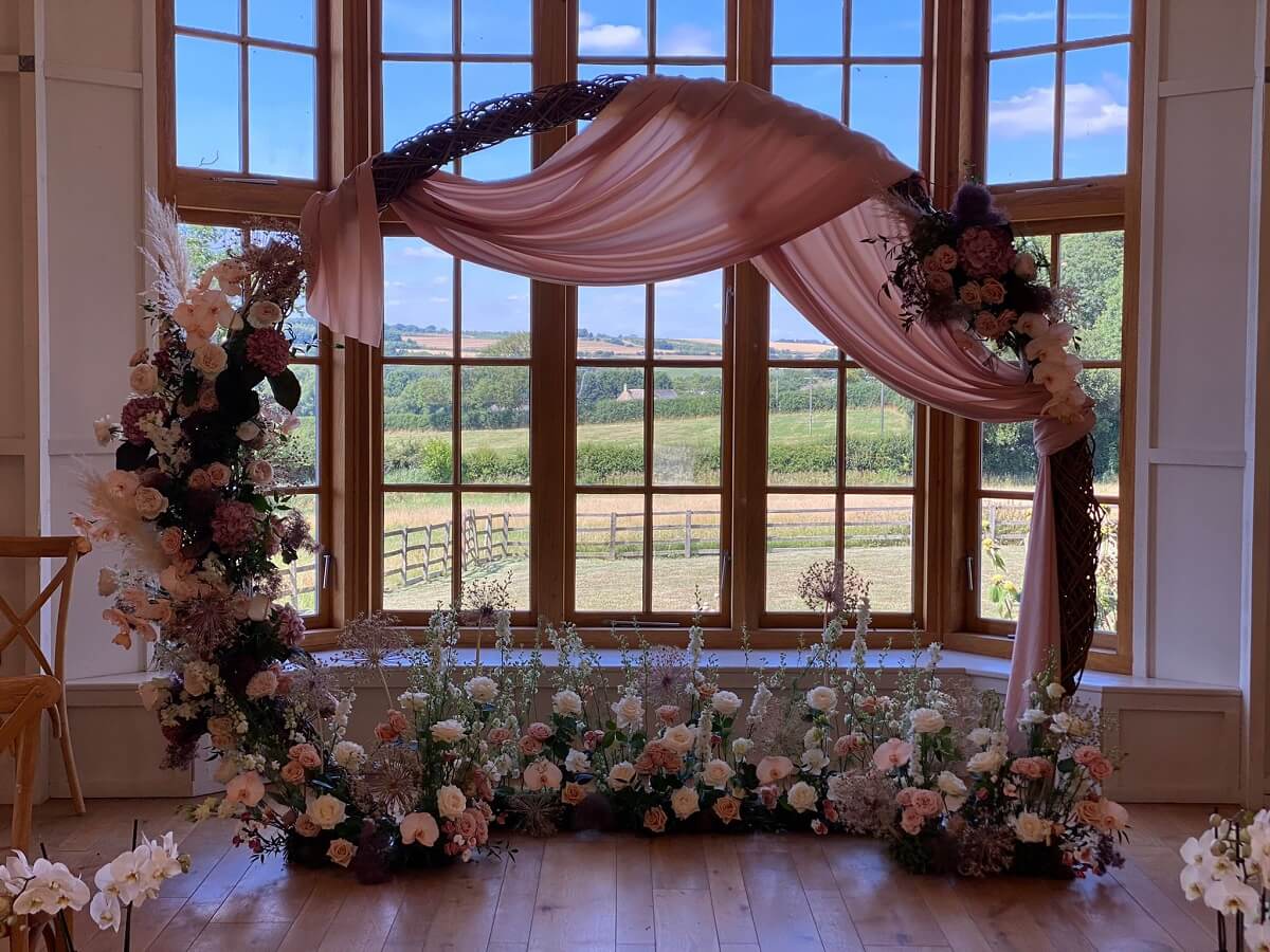 A large decorative moon arch formed with twisted canes, draped in salmon pink fabric, and decorated with white and pink flowers including roses, orchids, delphiniums, hydrangeas, carnations and more